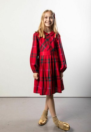 Młoda dziewczyna uśmiechająca się, ubrana w czerwoną sukienkę w kratę, złote buty z beżowymi skarpetkami, stojąca ze skrzyżowanymi nogami na jasnoszarej podłodze na białym tle.