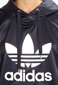 Marinblå huvtröja i slät tyg med en stor vit trefoil-logotyp och ordet "adidas" i fet vit text. Silverfärgade öglor.