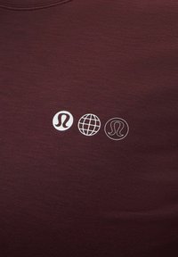 Három fehér logó, köztük két stilizált "L" alak és egy földgömb, egymás mellett nyomtatva sötét bordó szövetre.