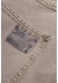 Țesătură din bumbac bej cu o etichetă cusută ce afișează "Wood Wood Copenhagen 2002" și logo-uri, având o textură fină de rigă și o strălucire subtilă.