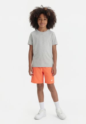 Dziecko z kręconymi włosami stoi, ma na sobie jasnoszarą koszulkę z krótkim rękawem, jaskrawo pomarańczowe spodenki z białym logo Nike, białe skarpetki i białe trampki.