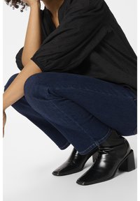 Personne portant un jean bleu foncé, des bottines noires à talons et un chemisier noir ample, accroupie avec une main près du visage.