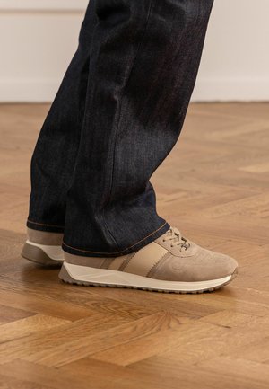 Feet wearing beige suede sneakers and dark blue jeans standing on a wooden parquet floor.