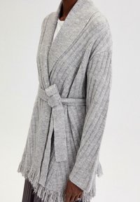 Šedý pletený kardigan s žebrovanou texturou, zavinovacím designem, páskem na zavázání a třásněmi na lemu. Vyroben z měkké látky, která zajišťuje pohodlí a teplo.