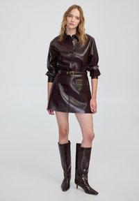 Conjunto de camisa y mini falda de cuero sintético burdeos con cintura ceñida. Botas de caña alta y puntera puntiaguda con acabado brillante. Textura suave.