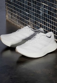 Ζευγάρι λευκά παπούτσια τρεξίματος Adidas Adizero με κορδόνια, τοποθετημένα σε σκοτεινό πάτωμα μπροστά από μπλε τοιχοπλακίδια.