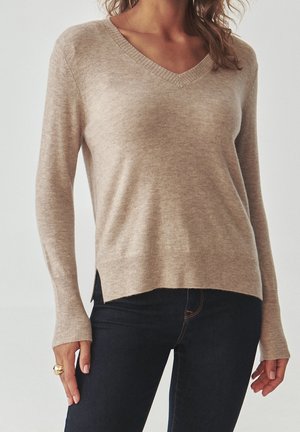 Žena v béžovém svetru s výstřihem do V a dlouhými rukávy a v černých džínách, stojící s rukama volně podél těla.