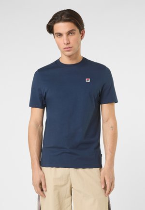 Giovane uomo con capelli castani corti che indossa una maglietta FILA blu scuro e pantaloncini beige su uno sfondo chiaro uniforme.