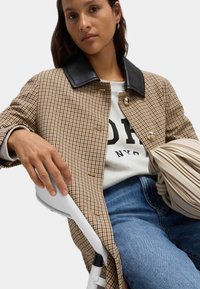 Cappotto scozzese con colletto nero, bottoni argentati, maglione grigio e jeans blu. Una borsa chiara è tenuta in una mano.