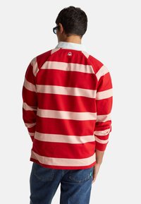 Mężczyzna ubrany w czerwono-różowy pasiasty, długi rękaw koszulki rugby z białym kołnierzykiem i niebieskie dżinsy, stojący tyłem do aparatu.