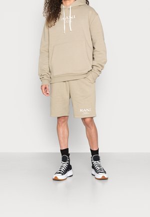 Beżowa bluza z kapturem ze sznurkami i przednią kieszenią, w zestawie z beżowymi szortami. Na szortach widoczne logo KANI. Czarne, wysokie trampki z białą podeszwą.