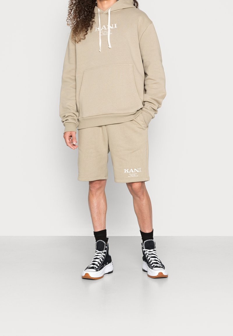 Béžová mikina s kapucí se stahovacími šňůrkami a přední kapsou, doplněná béžovými šortkami. Na šortkách výrazné logo KANI. Černé kotníkové tenisky s bílou podrážkou.