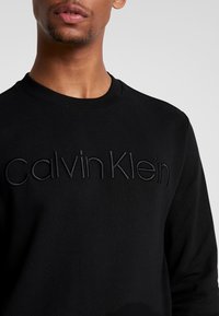 Felpa nera con logo ricamato 'Calvin Klein' in filo coordinato. Caratterizzata da collo a coste e texture del tessuto morbida.