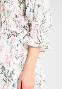 Vit blommig klänning i texturerad tyg, med rosa och blå blommönster och volangärmar med elastisk halsringning.