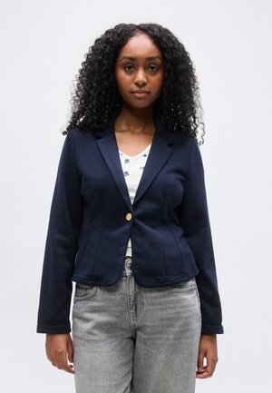 Ung kvinde med krøllet sort hår iført navy blazer over hvidt mønstret top og lysegrå jeans, stående mod en ensfarvet baggrund.