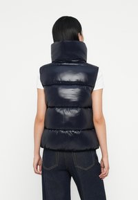 Marineblauw puffy vest met hoge kraag, glanzende afwerking en gesegmenteerde padding, gedragen over een wit shirt. Van achteraf weergegeven.