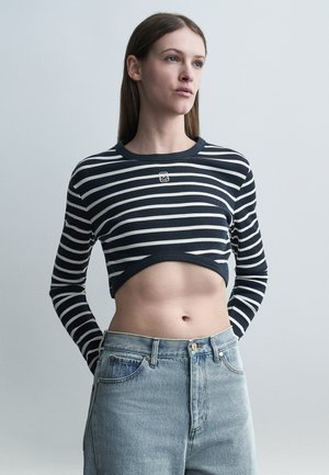 Mladá žena, ktorá má na sebe čierno-biely pruhovaný krátky sveter a svetlomodré rifle s vysokým pásom, stojaca pred jednoduchým sivým pozadím.