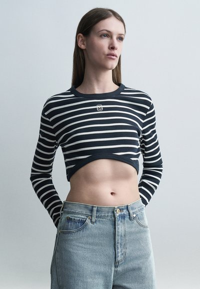 Young woman wearing a black and white striped cropped sweater and light blue high-waisted jeans, standing against a plain gray background.