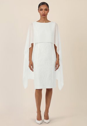 Mujer de pie vistiendo un vestido blanco texturizado hasta la rodilla con una capa translúcida superpuesta y tacones blancos de punta en un fondo beige.