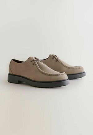Taupe suede shoes with a rounded toe, black rubber sole, and decorative stitching. Features thin laces and a smooth texture.