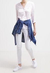 Bílá košile s knoflíky a červenými tečkami, bílé úzké džíny a uvázaná modrá denimová bunda. Celý outfit doplňují bílé tenisky.