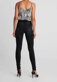 Jeans skinny negros combinados con una blusa de estampado metálico de serpiente, con tirantes finos y un corte holgado, vista desde la parte posterior con zapatos de tacón alto.