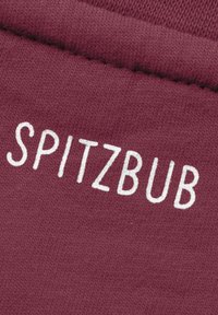 Bordeauxfarbener Stoff mit glatter Textur zeigt den weißen Text "SPITZBUB" in einer fettgedruckten, lässigen Schriftart.