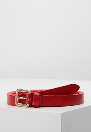 Rode leren riem met een kleine gouden metalen gesp, opgerold en liggend op een witte ondergrond tegen een effen lichtgrijze achtergrond.