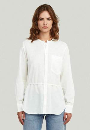 Femme aux cheveux bruns ondulés portant une chemise blanche à manches longues boutonnée avec une poche poitrine et un jean bleu clair, debout devant un fond uni.