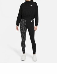 Czarna krótka bluza z kapturem z kieszenią z przodu, w zestawie z czarnymi legginsami z wzorzystą boczną wstawką. Biały logo Nike na obu elementach. Białe sneakersy.