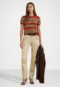 Haut en tricot avec des motifs rouges, verts et jaunes ; pantalon cargo beige avec zips sur le côté ; veste en cuir marron ; sandales à talons hauts noires.