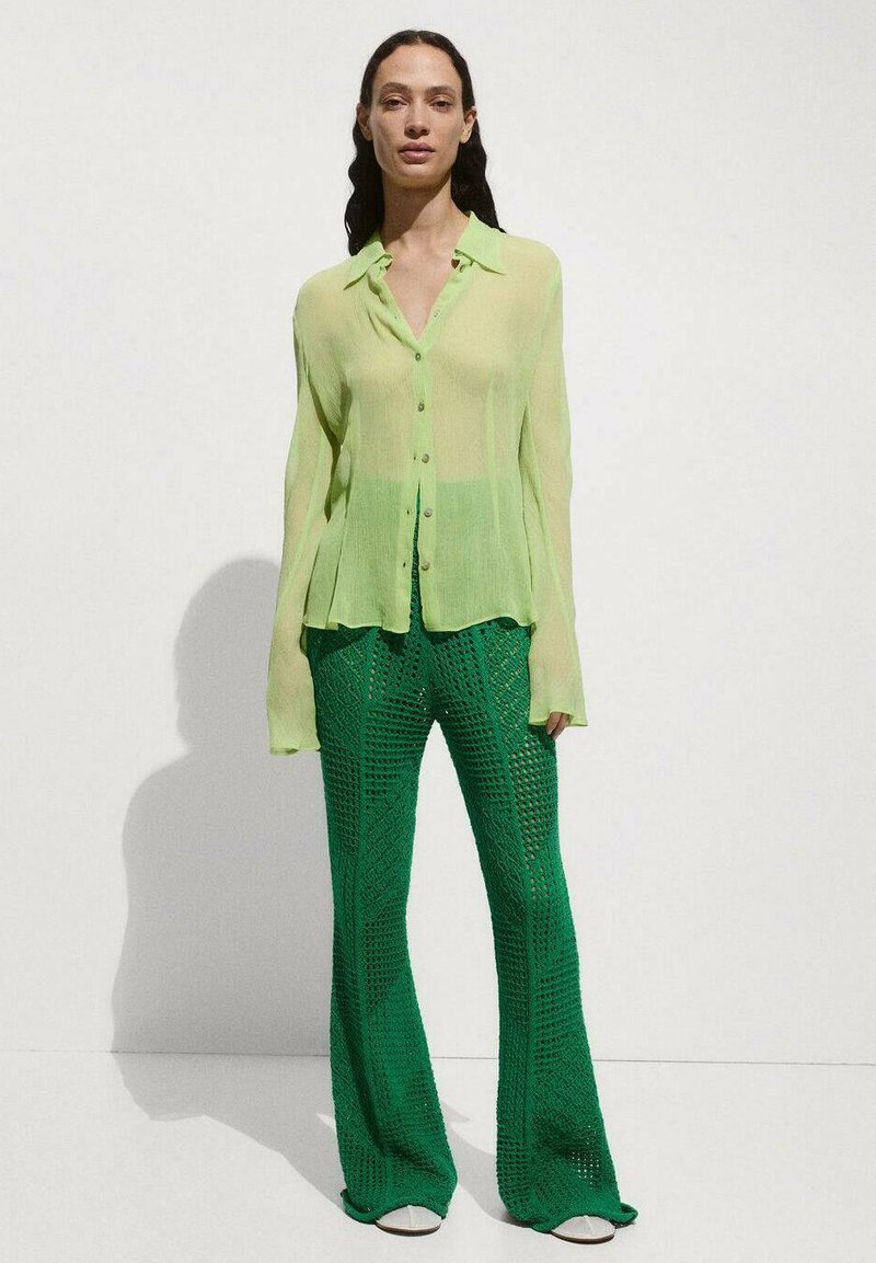 Camicia verde chiaro trasparente con colletto, abbinata a pantaloni larghi verdi con una trama strutturata e motivo a rombi. Calzature bianche visibili.