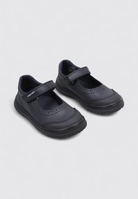 Par de zapatos infantiles negros estilo Mary Jane con correas de Velcro y suelas de goma texturizadas sobre un fondo claro.