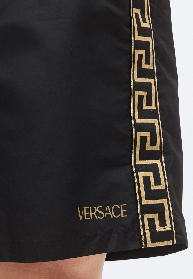 Shorts noirs Versace avec logo doré et motif clé grecque sur le côté, réalisés en tissu lisse et léger.