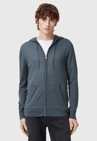 Felpa con cappuccio e zip in verde scuro, con tasche frontali, polsini a costine e una texture liscia. Indossata sopra una maglietta bianca.