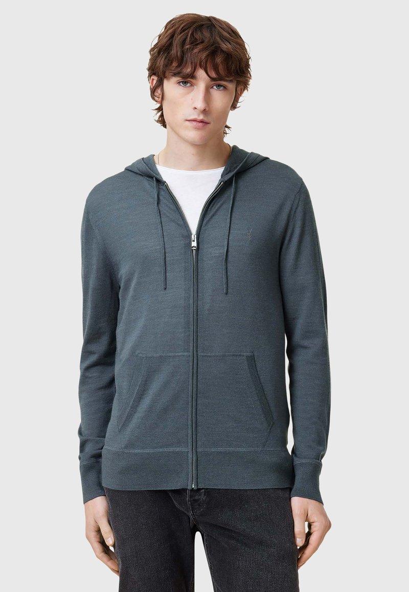 Felpa con cappuccio e zip in verde scuro, con tasche frontali, polsini a costine e una texture liscia. Indossata sopra una maglietta bianca.