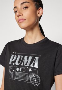 T-shirt noir avec un design imprimé argenté comportant le mot "PUMA", des formes géométriques et des accents sur un tissu lisse.