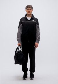 Μαύρο και γκρι αθλητικό σετ με ζακέτα με φερμουάρ, ελαστικά μανσέτα και αντίστοιχο παντελόνι· κρατά μια μαύρη τσάντα τύπου duffel. Μαύρα αθλητικά παπούτσια.