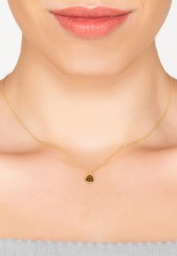 Collar de cadena de oro con un pequeño colgante de piedra verde en forma de gota. La cadena presenta una textura retorcida, reposando sobre piel desnuda.