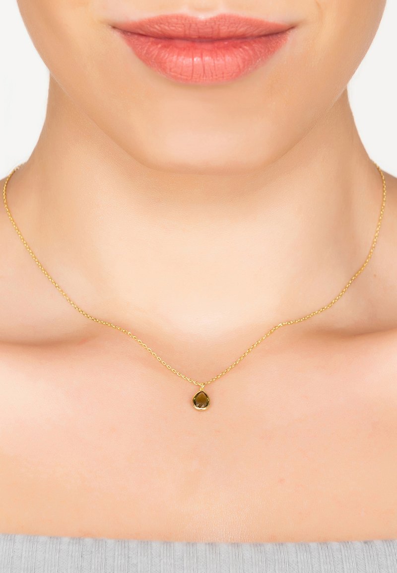 Collar de cadena de oro con un pequeño colgante de piedra verde en forma de gota. La cadena presenta una textura retorcida, reposando sobre piel desnuda.