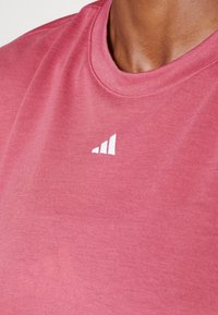 Różowa koszulka z krótkim rękawem i okrągłym dekoltem, wykonana z miękkiego materiału, z białym logo Adidas na przodzie. Gładka faktura z delikatnym połyskiem.