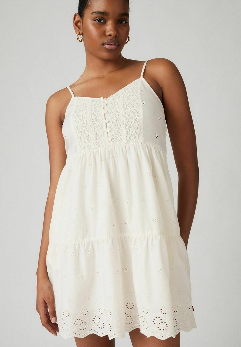 Vestido de algodón blanco con tirantes ajustables, que presenta patrones de ojal bordados, un cierre frontal con botones, una falda en capas y un dobladillo en forma de onda.