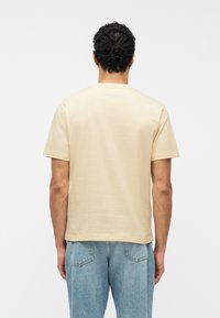 Άνδρας με σγουρά σκούρα μαλλιά που φοράει απλό μπεζ κοντομάνικο μπλουζάκι και μπλε τζιν, γυρισμένος με την πλάτη σε λευκό φόντο.