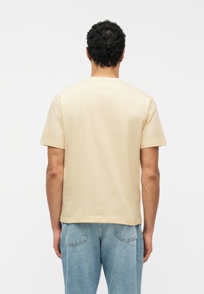 Homme aux cheveux bouclés foncés portant un t-shirt beige uni à manches courtes et un jean bleu, vu de dos sur un fond blanc.