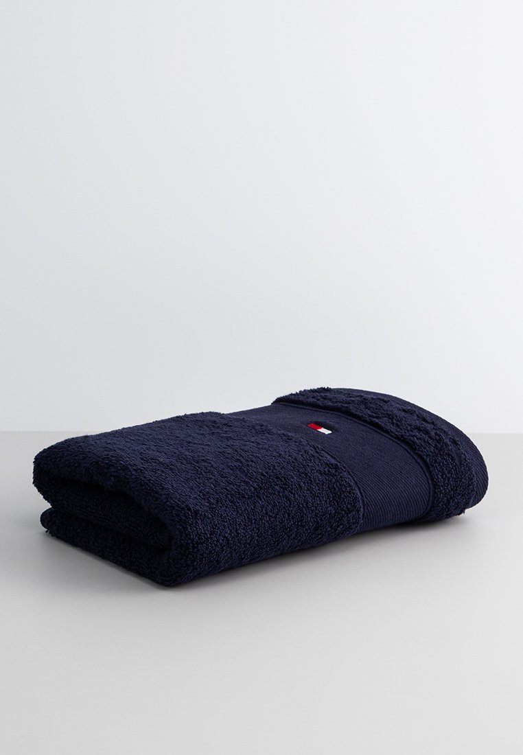 Serviette bleu marine en coton, avec une surface texturée et une bande lisse, ornée d'un petit logo rouge, blanc et bleu.