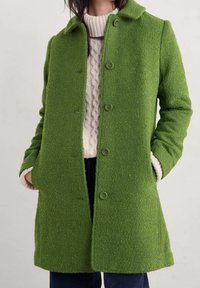 Zielony wełniany płaszcz o fakturowanym wykończeniu, z zapinaniem na guziki z przodu i bocznymi kieszeniami. Posiada klasyczny kołnierz i jest zestawiony z kremowym swetrem pod spodem.