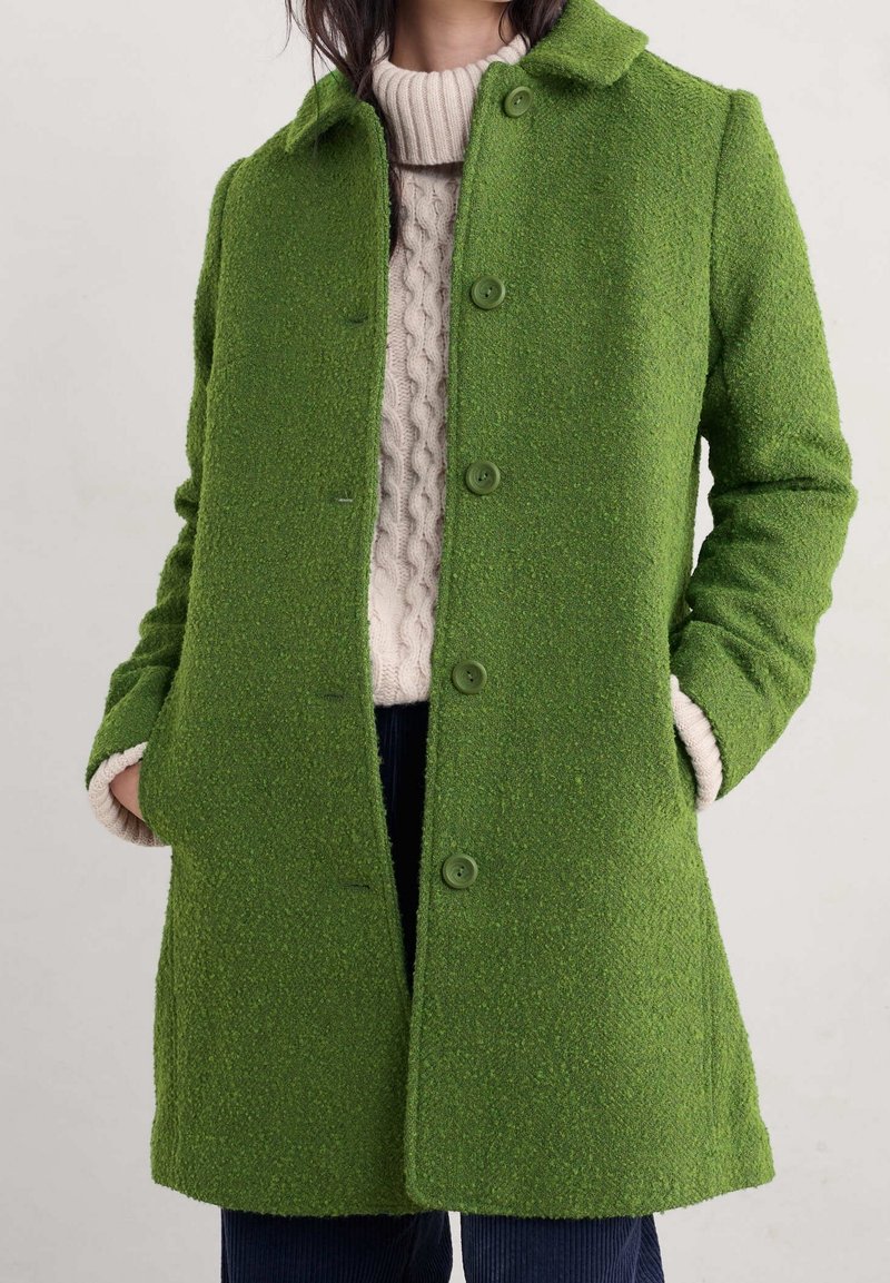 Zielony wełniany płaszcz o fakturowanym wykończeniu, z zapinaniem na guziki z przodu i bocznymi kieszeniami. Posiada klasyczny kołnierz i jest zestawiony z kremowym swetrem pod spodem.