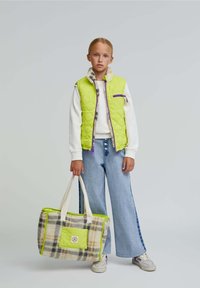 Chaleco acolchado verde lima con cremallera morada, suéter blanco y pantalones de mezclilla acampanados azul claro. Bolsa de mano de cuadros beige con acentos y asas verdes.