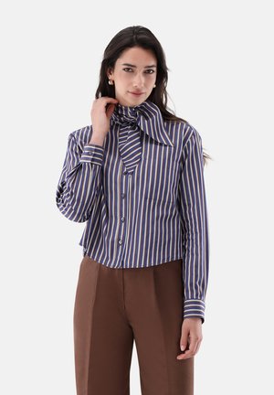 Frau trägt eine blau-weiß-braun vertikal gestreifte Bluse mit einer Schleife am Kragen und braune, hoch taillierte Hosen, stehend vor einem einfarbigen Hintergrund.