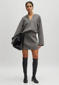 Grauer oversized Pullover mit V-Ausschnitt und langen Ärmeln, kombiniert mit einem enganliegenden grauen Minirock und hohen schwarzen Lederstiefeln. Hält eine schwarze Tasche.